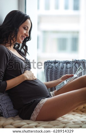 Side view of happy young pregnant woman looking at ultrasound scan on window sill