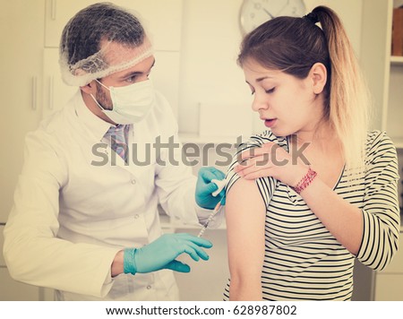 Smiling man doctor making injection of vaccine to teenager girl patient in hospital