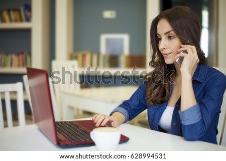 Pretty brunette women sitting at the table and working at a laptop and speaking on smartphone. Co working zone. Pretty freelancer.