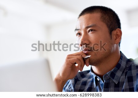 Young Asian designer deep in thought with a hand on his chin while working on a laptop alone at a desk in a modern office Royalty-Free Stock Photo #628998383