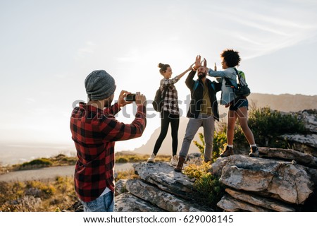 Young man taking pictures of his friends doing high five in countryside while hiking. Group of hikers enjoying during summer vacation.