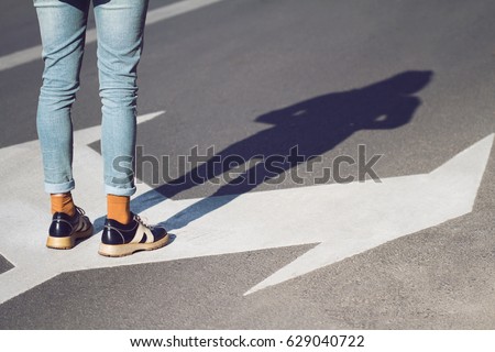 side view close up of a young woman wearing black shoes and blue jeans standing on a street with arrow signs pointing in different directions concept for life choices Royalty-Free Stock Photo #629040722