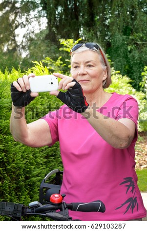 Senior woman on a bicycle tour in summer wearing protective goves, using her cell phone to take pictures of the landscape, green background
