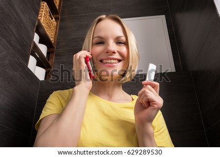 A beautiful young blonde woman sits in the bathroom and holds a positive pregnancy test and red mobile phone. Conceptual photography.