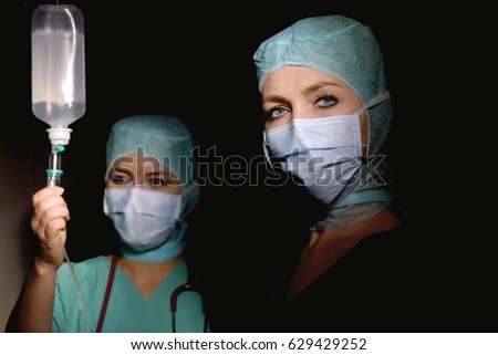 Two young women pose in a low lit operation theater. 
Fully dressed as theater nurses with face masks 
and green sterile medical work clothing.
