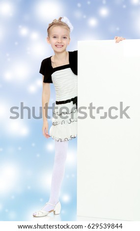 Beautiful little blonde girl dressed in a white short dress with black sleeves and a black belt.The girl peeks out from behind white banner.Blue Christmas festive background with white snowflakes.