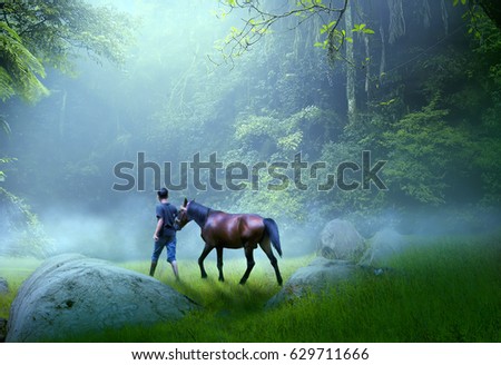 A man with a horse walking on the grass on the edge of a dark forest with a light that breaks through between the trees and the background of the natural misty forest