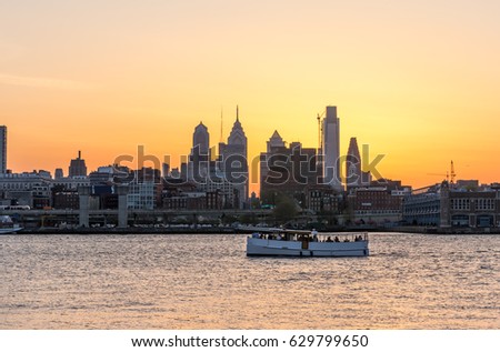 Skyline in Philadelphia Pennsylvania at sunset against a bright orange sky with a boat in the foreground traveling down the Delaware River.