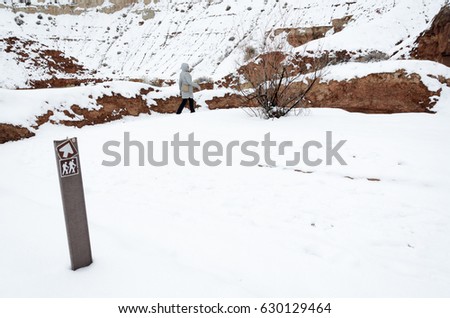 Hiking trail sign in the foreground, hiker walking at winter on trail in the background. Arrow shows direction. Hiking in extreme weather conditions, follow the path, stay in bounds after snow storm.