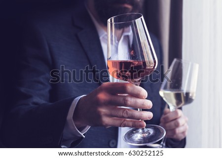 horizontal close up of a Caucasian man with beard black suit and white shirt offering a tall glass of rose wine at an event by the window natural light Royalty-Free Stock Photo #630252125