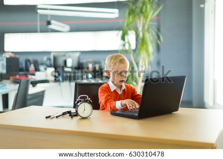 Young blonde caucasian kid in orange shirt sit at the table with colorful folders and alarm clock and plays on the black laptop
Blurred office on background