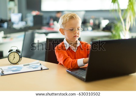 Young blonde caucasian kid in orange shirt sit at the table with colorful folders and alarm clock and plays on the black laptop, look at laptop
Blurred office on background