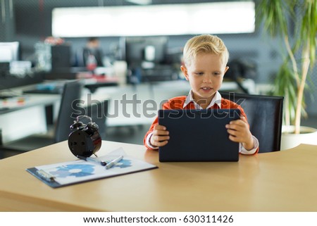 Young blonde caucasian kid in orange shirt sit at the table with colorful folders and alarm clock and plays on the black tablet, amased, glad
Blurred office on background