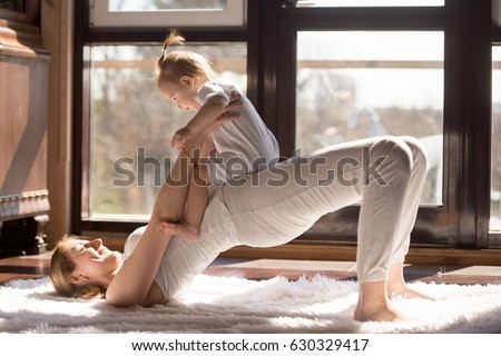 Young yogi smiling mother doing half bridge exercise, wearing white sportswear, working out with baby daughter, family yoga at home, having no time for sport club. Healthy lifestyle concept photo  Royalty-Free Stock Photo #630329417