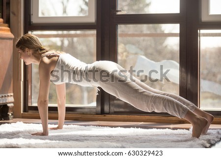 Young yogi attractive woman practicing yoga concept, standing in Push ups or press ups exercise, Plank pose, phalankasana, working out, wearing white sportswear, full length, home interior background 