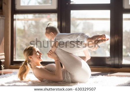 Young sporty mother and baby girl working out, exercising together at home, parent and child healthy development, game playing, fitness and relaxation, wearing white. Healthy lifestyle concept photo  Royalty-Free Stock Photo #630329507