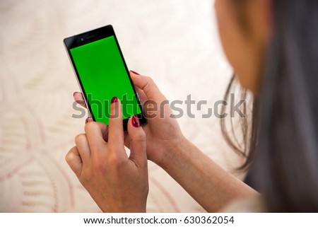 Young woman types and slides on her smartphone at home. Cellphone with chroma key screen - green screen. Red nail polish fingers.