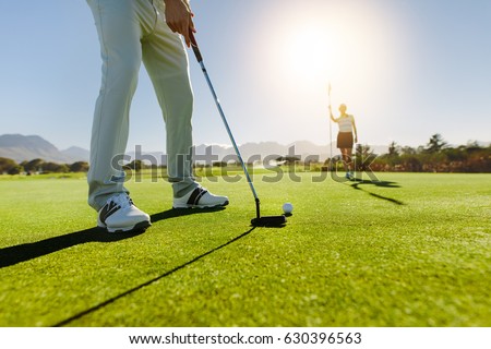 Low angle view of golfer on putting green about to take the shot. Male golf player putting on green with second female player in the background holding the flag.