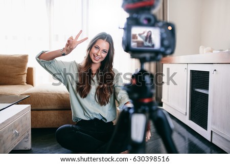 Young woman vlogger showing victory or peace sign while recording her daily video blog. Vlogger using a camera mounted on a tripod to record her video.