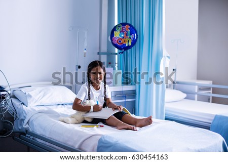 Smiling girl drawing picture in a book while sitting on hospital bed 