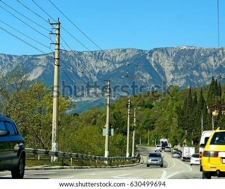 Cars are driving along a road that curves, along the roadside are green trees, poles, wires for trolley buses, in the background are visible blue mountains and sky