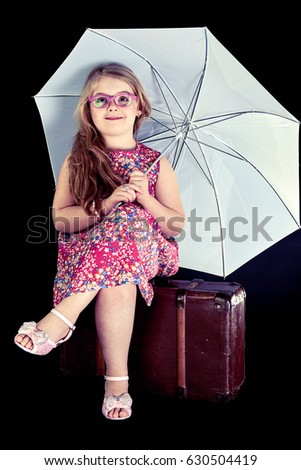 Portrait of funny adorable little blonde girl playing with an umbrella, sitting on a old suitcase, Wearing a nice floral dress,isolated on black background