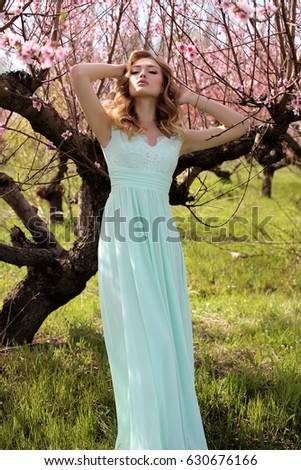 fashion outdoor photo of gorgeous young woman with blond hair in elegant dress posing in garden with blossom peach trees
