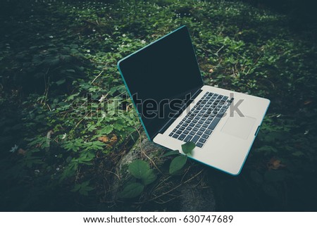 Freelancer laptop computer lost in the forest glade, amid the greenery, summertime. Photo depicts notebook computer in the woods, blurred forest green plants behind. Freelance concept.