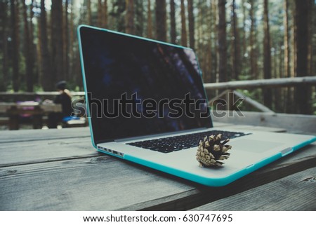 Freelancer laptop computer in the forest on the wooden textured table with cone, summertime. Photo depicts notebook computer in the woods, blurred forest green trees behind. Freelance work concept.