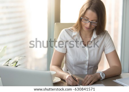 Satisfied businesswoman signs a contract at workplace. Young woman entrepreneur sitting at the desk and writing business letter. Female office head puts personal signature on company agreement 