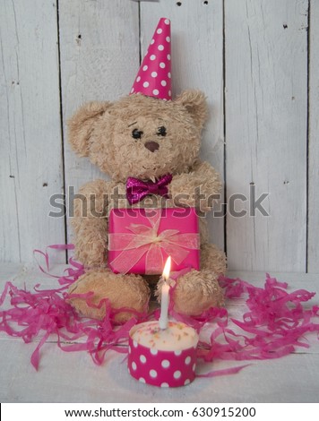 Cute tatty teddy bear wearing a pink party hat and pink bow tie holding a wrapped birthday present with a birthday cake and lit candle 