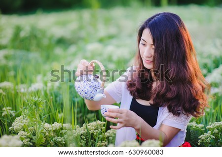 Young Chinese smiling girl pouring tea outdoors. Asian beauty posing for photos, holding small tea pot. Pretty girl sitting in flowering meadow, grass and herbs, lit with sunset rays of light.