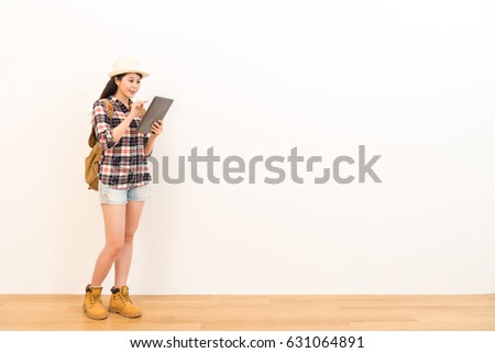 smiling female backpacker using digital tablet computer searching tourist destination guidebook through internet standing on wooden floor with white wall background.