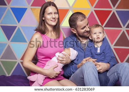 Cute caucasian ordinary family in casual clothes at home sitting on a couch. Lifestyle portrait of mom, dad, two years old son and newborn daughter. Colorful background
