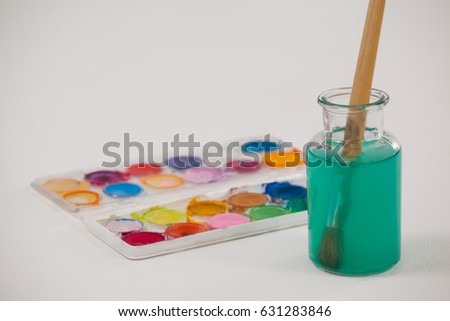 Watercolor palette and paintbrush with blue paint dipped into a jar filled with water against white background
