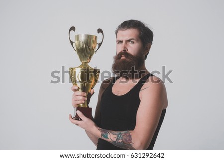 bearded man, long beard, brutal caucasian hipster with moustache in black vest holds gold champion cup with serious face on grey background, unshaven guy