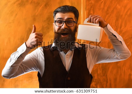 Bearded man, long beard. Brutal caucasian unshaven amused hipster with glasses and moustache wearing white shirt, suede waistcoat, holding meal box showing gesture yes on brown studio background