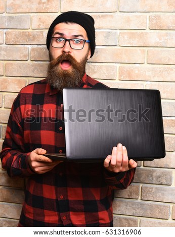 Bearded man, long beard. Brutal caucasian surprised unshaven hipster holding laptop in red black checkered shirt with hat and glasses on beige brick wall studio background