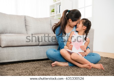 happy mom holding happy little daughter and giving her kiss sitting on the living room floor when she knowing girl prepare mother's day card gift.