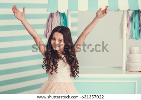 Beautiful school girl in dress is keeping hands raised, looking at camera and smiling, on light blue and pink background