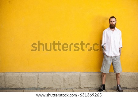 Tattooed bearded hipster on yellow wall background posing outdoor. Cool fashion man in summer shorts and white shirt. Colorful, trendy and fashionable guy