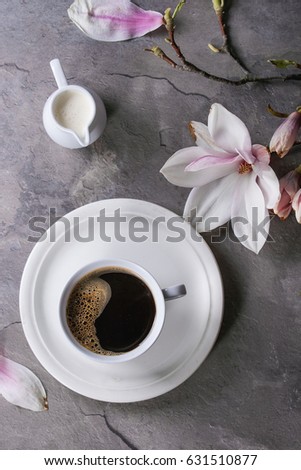 White cup of black coffee, served on white saucer with jug of cream and magnolia flower blossom branch over gray texture background. Flat lay, space