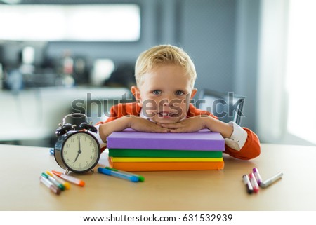 Cute blonde caucasian kid sit at the table with colorful folders and alarm clock and relies on the colorful folders, hands on the folder, look at camera
Blurred office background, focus on lad