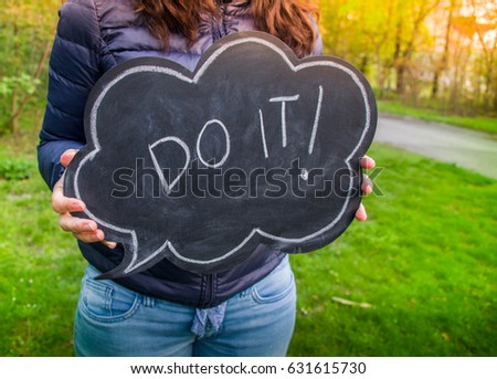 Do it text blackboard. Young woman holding a blackboard with speech bubble shape with the words do it written in English as a concept of taking action and motivation, on a natural background