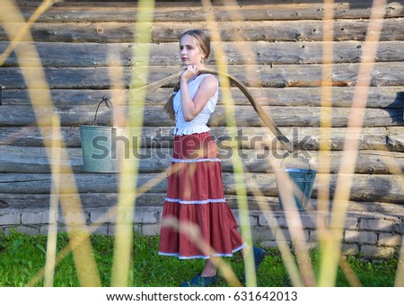 In the distance a young girl with a yoke and buckets on the background of a log house. The photo is taken through the grass.