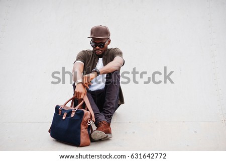 Portrait of sitting stylish african american man wear on sunglasses and cap with handbag outdoor. Street fashion black man.