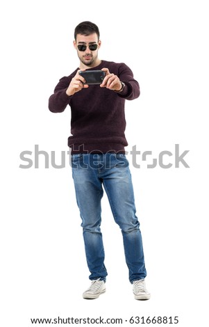 Cool young casual man recording video with mobile phone aiming at camera. Full body length portrait isolated over white studio background.