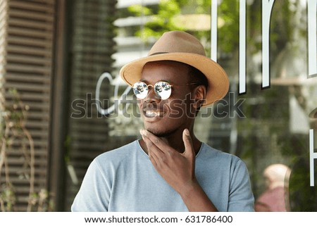 Cheerful young Afro American male in trendy sunglasses and headwear touching his chin and smiling happily as he sees his friend approaching him while waiting for lunch at sidewalk restaurant