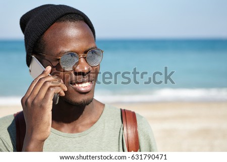 Handsome happy Afro American student wearing round shades and hat smiling broadly, talking on mobile phone to his parents, saying that he is doing ok while traveling alone during summer holidays