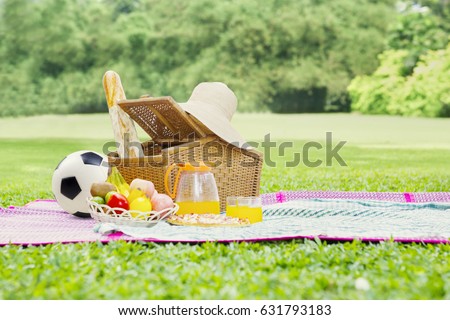 Picture of a picnic basket on mat with foods, drink, straw hat, and a soccer ball. Shot at green meadow